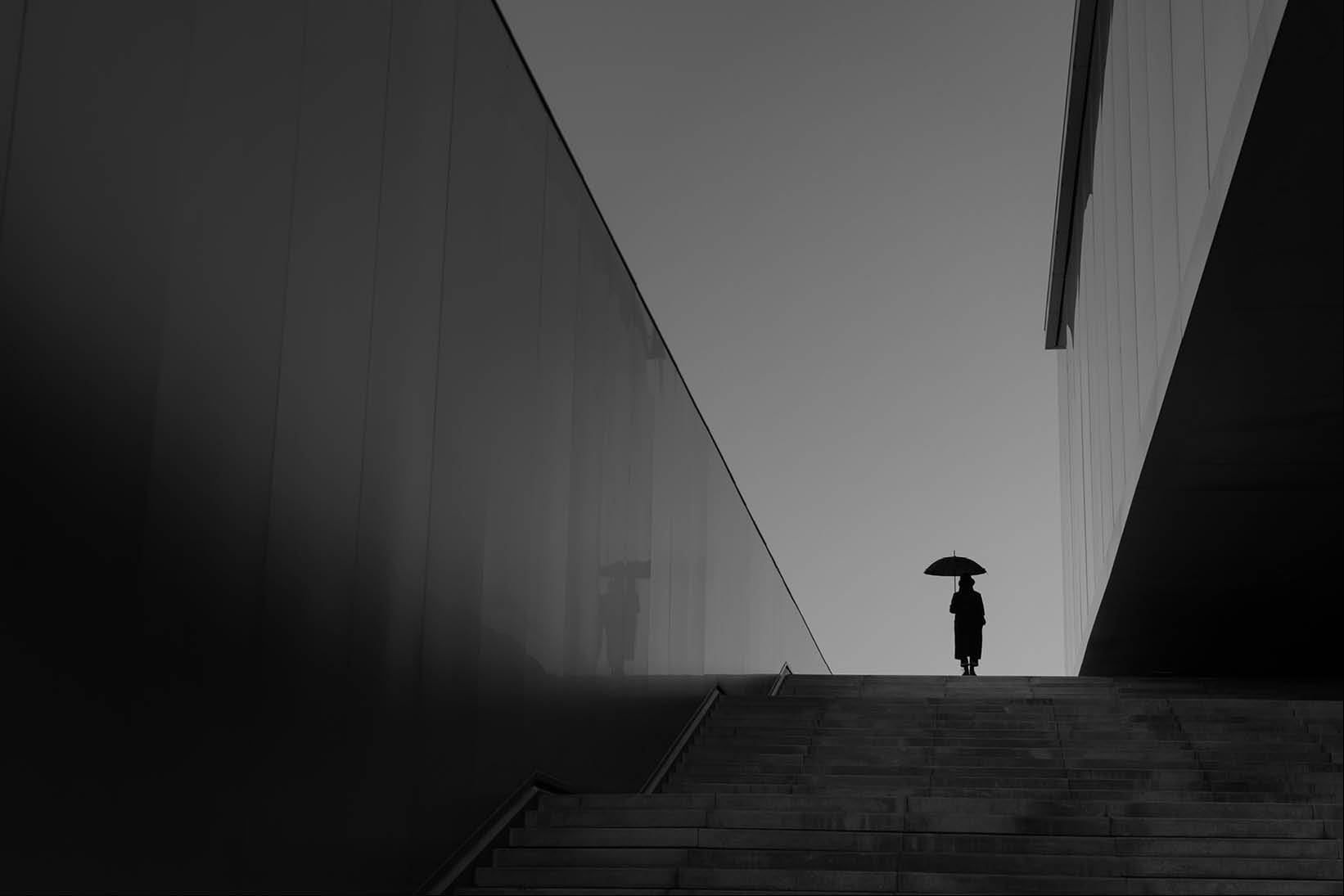 Silhouette of a person holding an umbrella at the top of a staircase between modern buildings, representing protection, resilience, and confidence in an uncertain environment - Used on Capco Insurance
