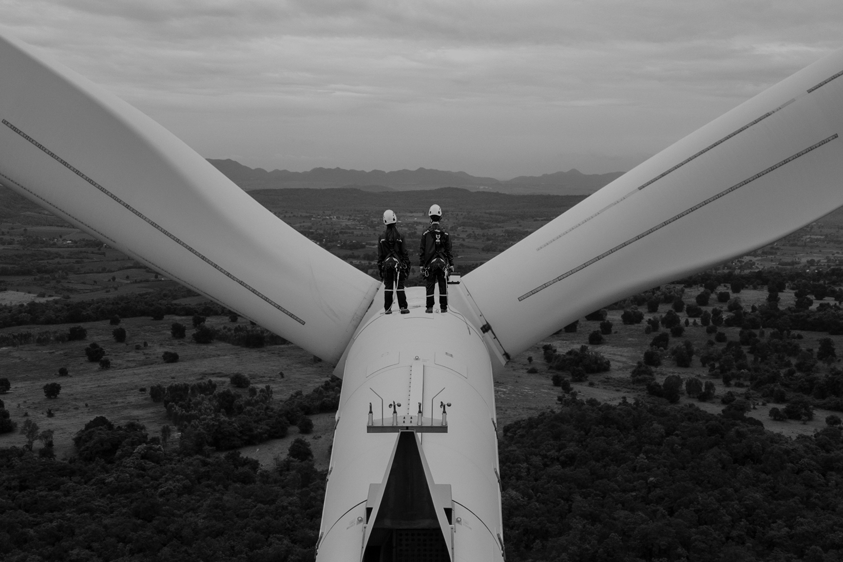 Two technicians standing atop a wind turbine overlooking a landscape with additional turbines.